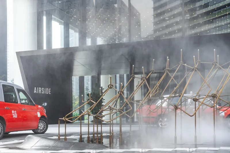 Brass geometric installation sculpture releasing mist in front of a dark concrete wall beneath a glass canopy