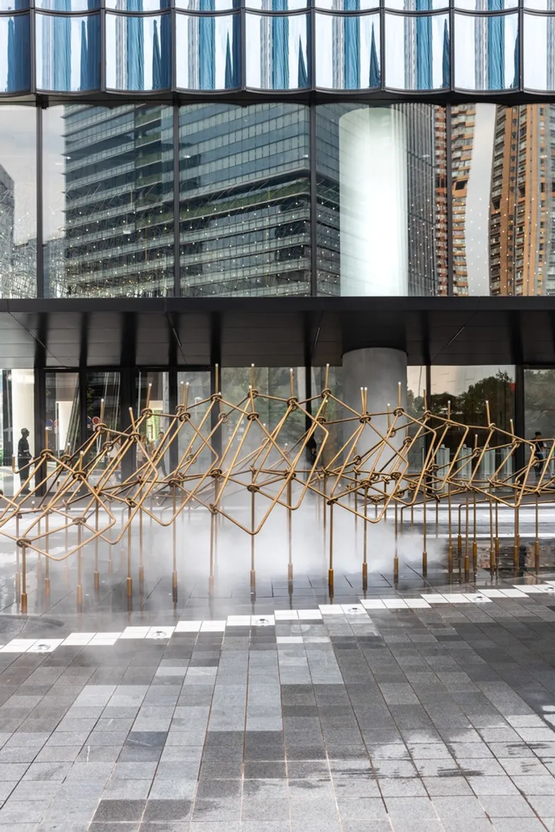 Angular brass rod sculpture on a paved plaza beneath a glazed pedestrian bridge in bright daylight
