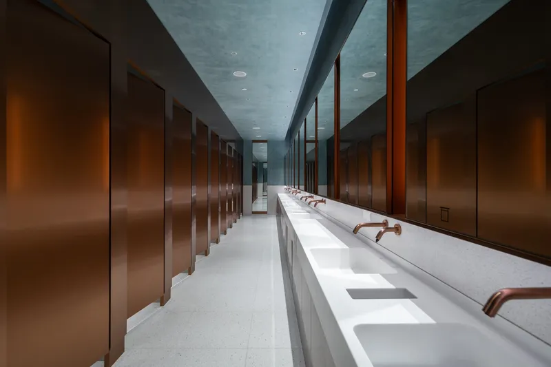 Restroom interior with bronze partition stalls and continuous counter with copper fixtures under a blue-grey ceiling