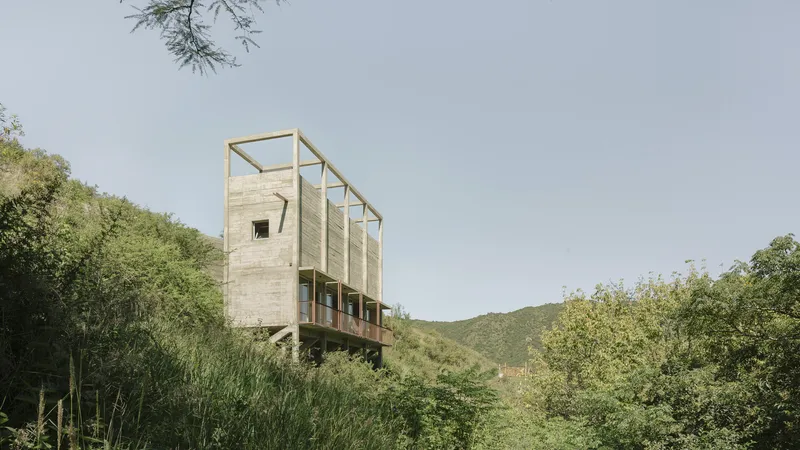 Elevated board-formed concrete volume on pilotis with open frame roof rising above dense hillside vegetation