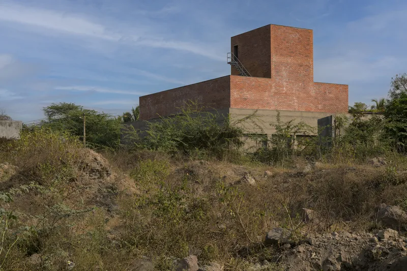 Red brick volumes rising above overgrown terrain with dry grasses and scattered trees
