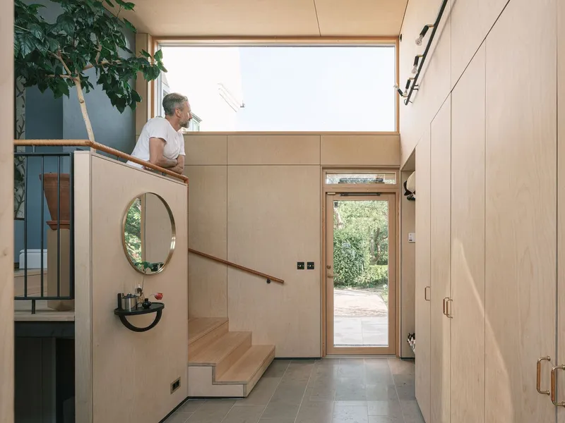 Entry hall with plywood-clad staircase and person ascending toward clerestory window above the glazed door