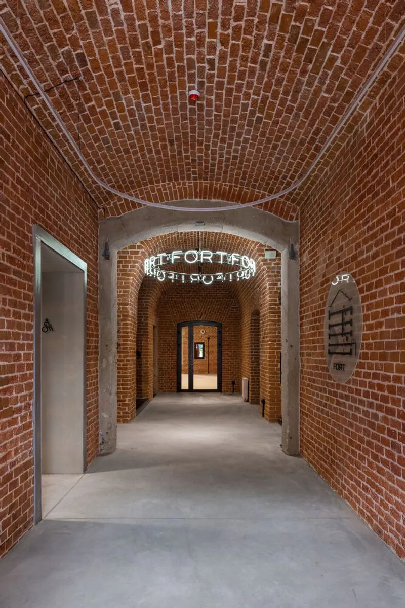 Barrel-vaulted brick corridor with neon signage and concrete floor leading to distant doorway