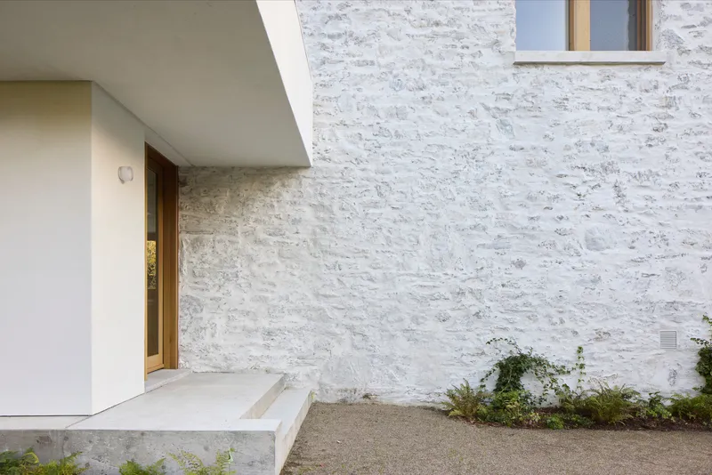 Recessed timber entrance door beneath concrete canopy beside whitewashed stone wall with small planting bed