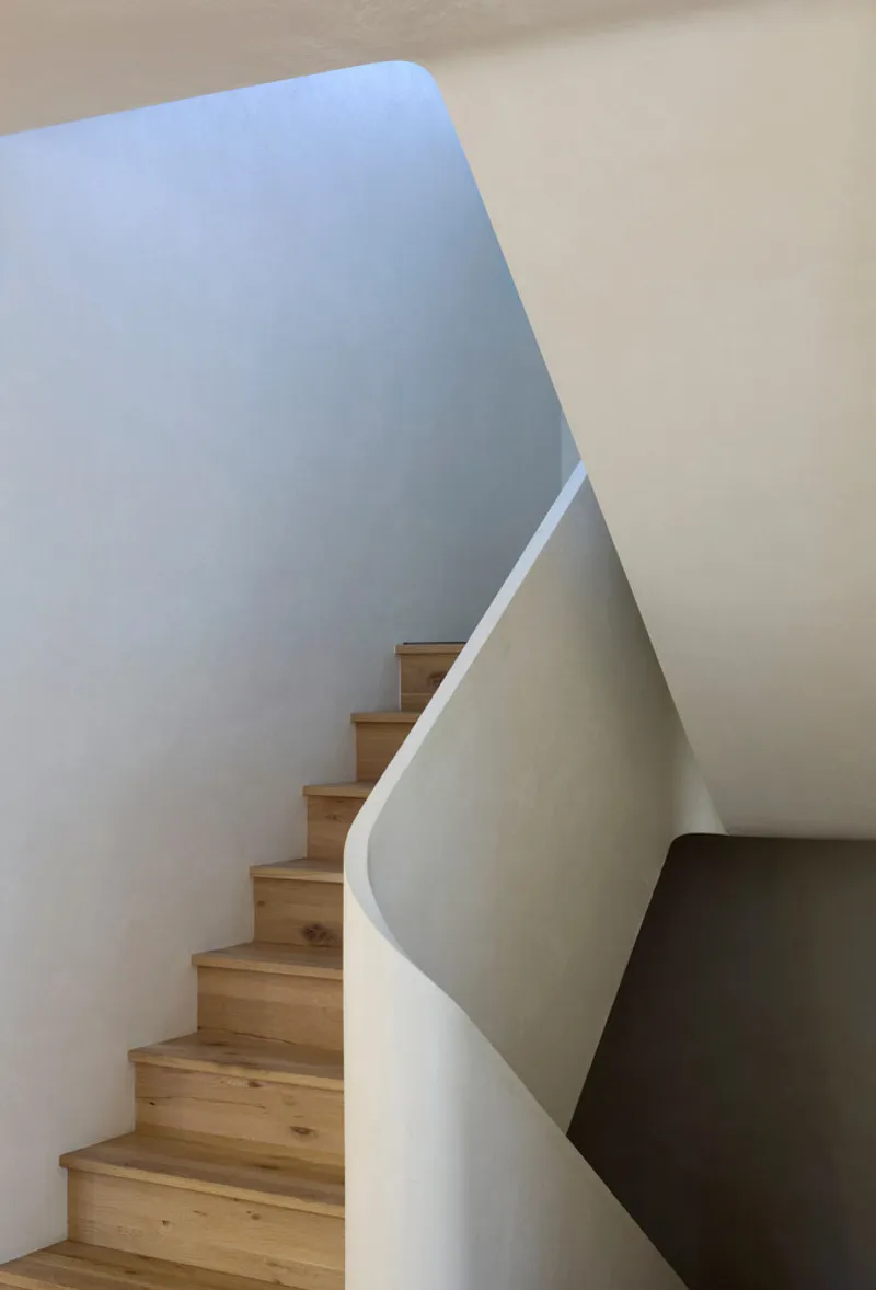Timber staircase ascending between curved white walls beneath a skylit opening with blue sky visible