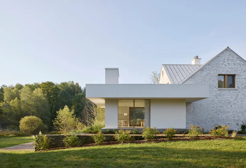 White rendered extension with deep overhang adjoining a whitewashed brick gable with metal roof in green landscape