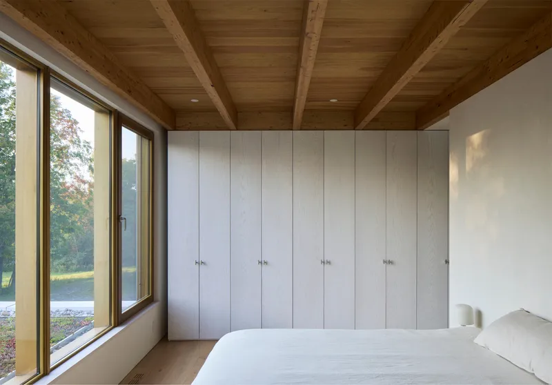 Bedroom with light oak wardrobes, exposed timber ceiling joists and full-height glazing overlooking lawn