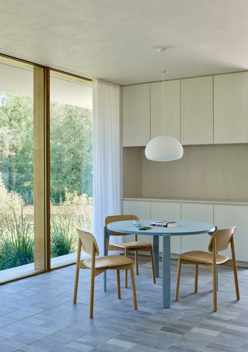 Dining area with pale green cabinetry, blue round table and floor-to-ceiling glazing overlooking planted garden