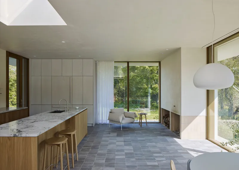 Open-plan kitchen and living space with grey slate tile floor and glass doors framing tree canopy beyond