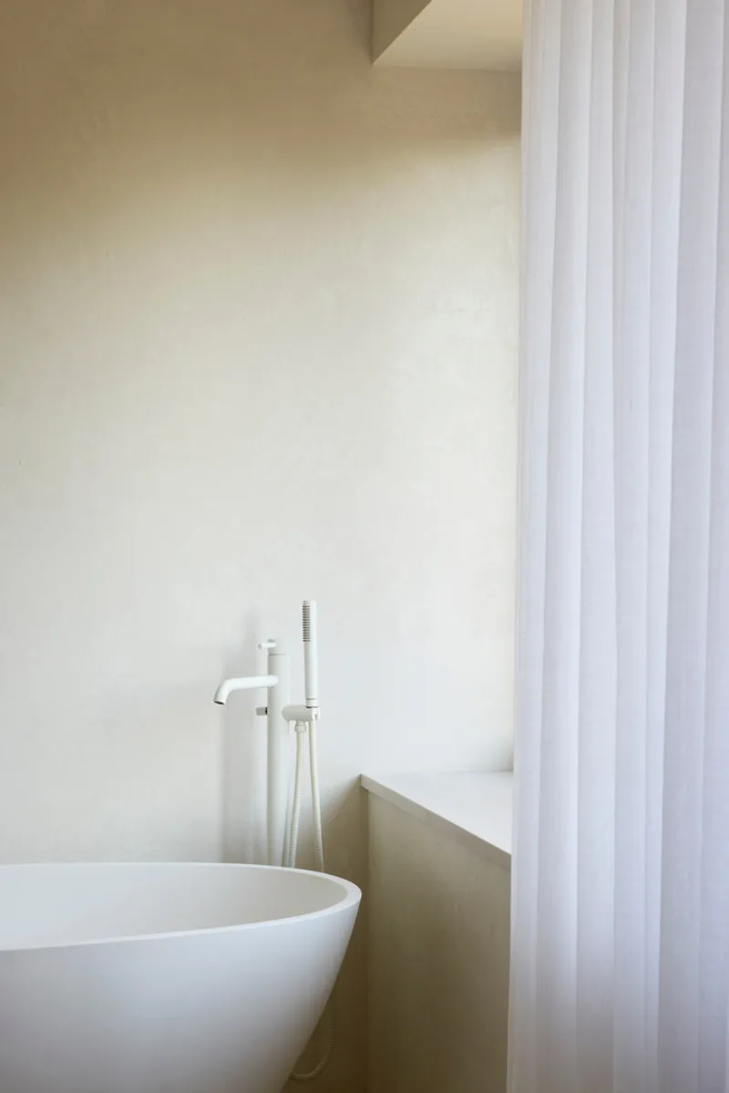 Freestanding white bathtub with floor-mounted tap beside vertical sheer curtain under clerestory light
