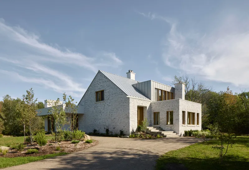 Front facade combining white-painted stone gable and new rectilinear volume with timber windows under blue sky
