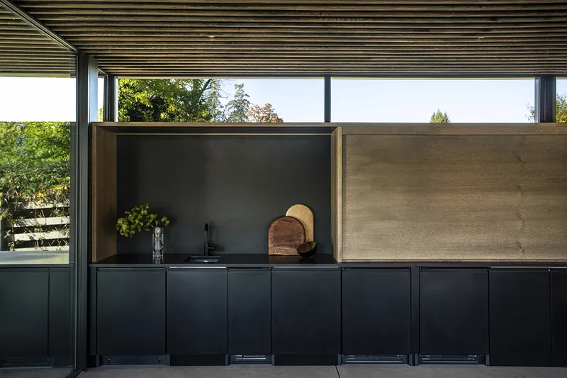 Outdoor kitchen with black cabinetry and wood backsplash beneath a slatted timber ceiling