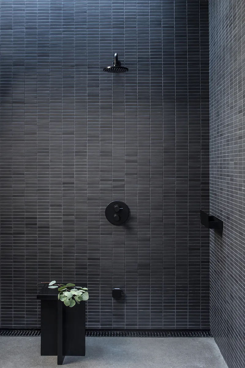 Shower wall clad in narrow horizontal dark grey tiles with black fixtures and plant stand