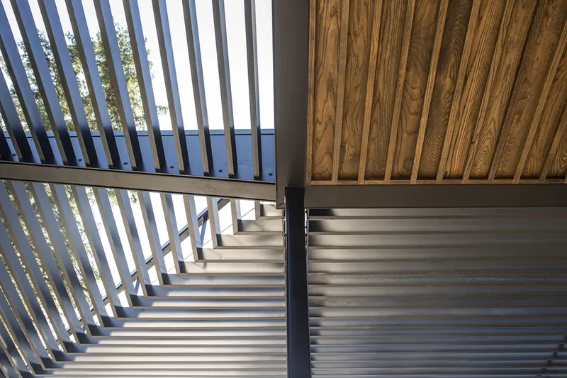 Staircase with vertical metal slat screen filtering daylight beneath a wood-panelled ceiling