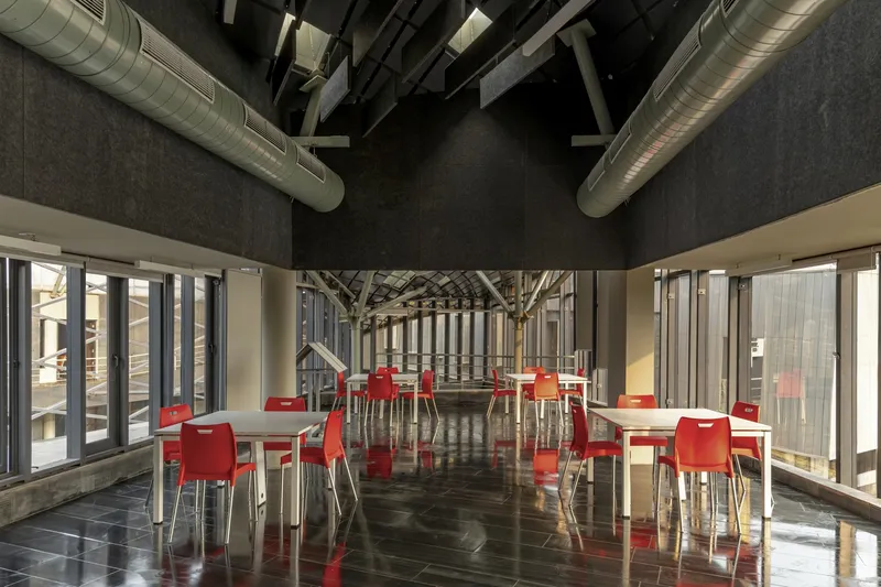 Interior dining space with red chairs, polished black floor, exposed ducts, and timber structure beyond glass walls