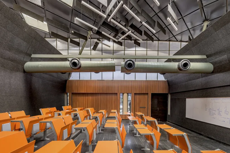 Lecture hall interior with orange seating under exposed cylindrical ventilation ducts and skylight