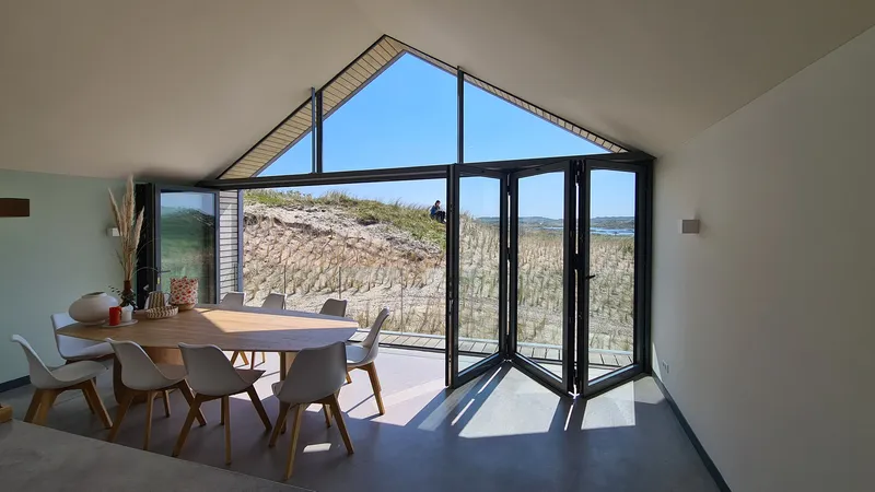 Interior dining space with folding glass doors opening to dune landscape framed by vaulted ceiling and clerestory
