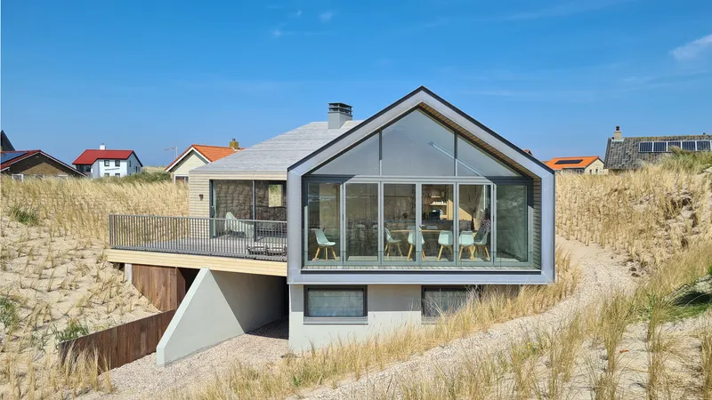 Gabled glazed facade with timber deck and metal railing embedded in dune vegetation between neighboring houses