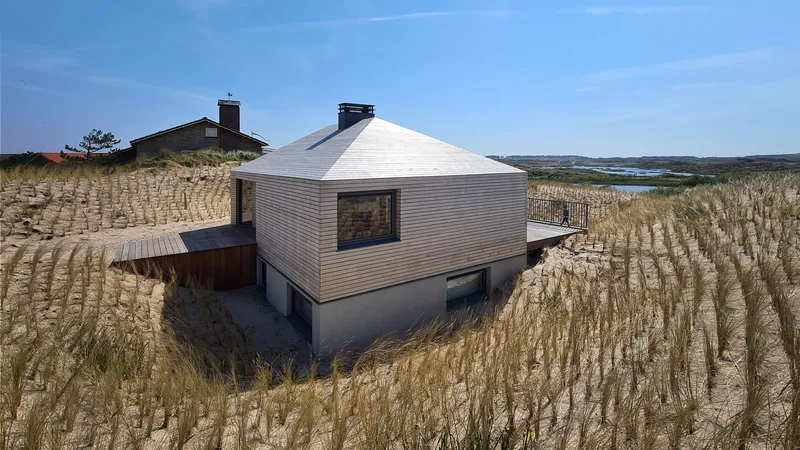 Weathered timber and white render volumes nestled in dune grasses with coastal bay visible beyond