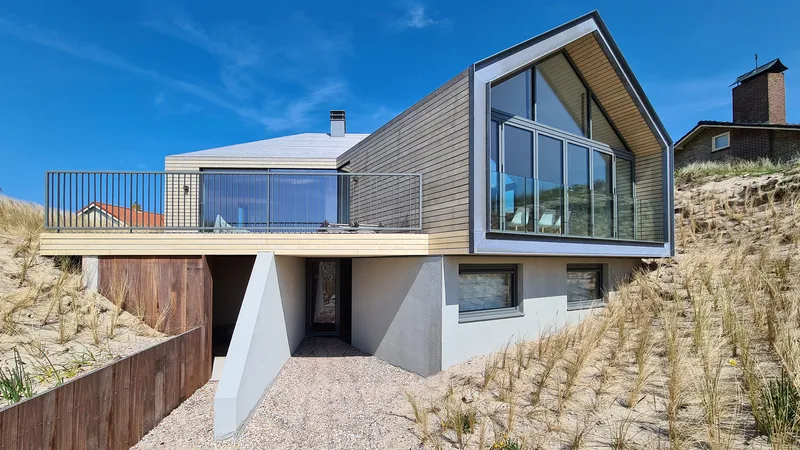 Angled facade with horizontal timber cladding and glazed gable end set into sandy hillside