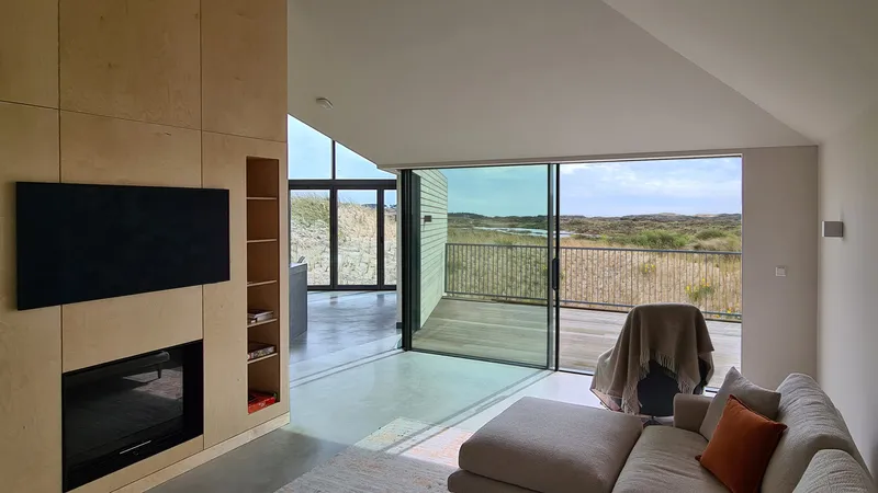 Living room with plywood wall unit and floor-to-ceiling glazing framing dune landscape views