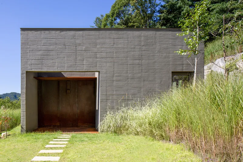 Horizontally striated concrete facade with recessed timber garage door and tall grasses under blue sky