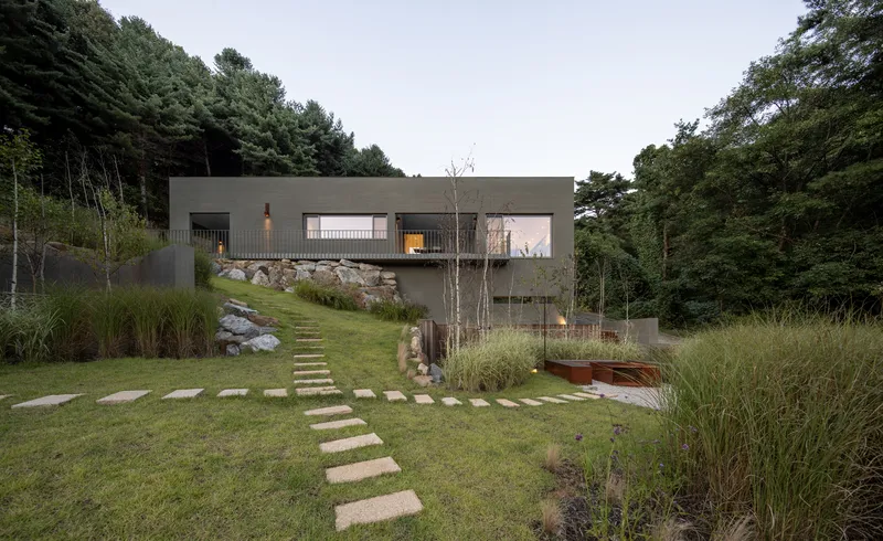 Stepped stone pathway leading through planted grasses to the elevated concrete volume at dusk