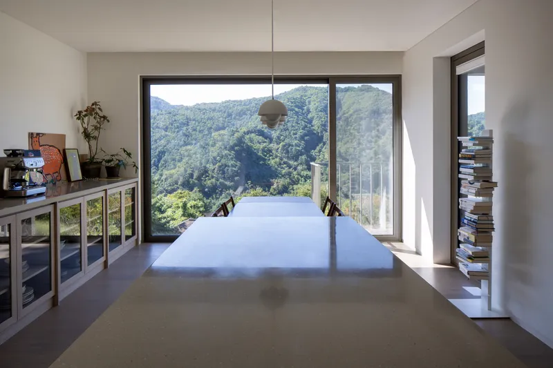 Long white table in workspace with pendant light and panoramic window framing forested mountain valley