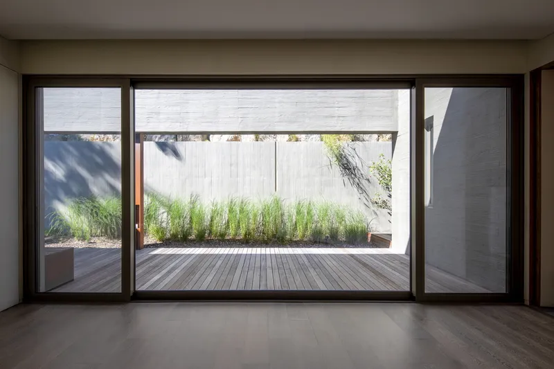 Interior view through full-height glazing to timber deck courtyard with planted bed of tall grasses