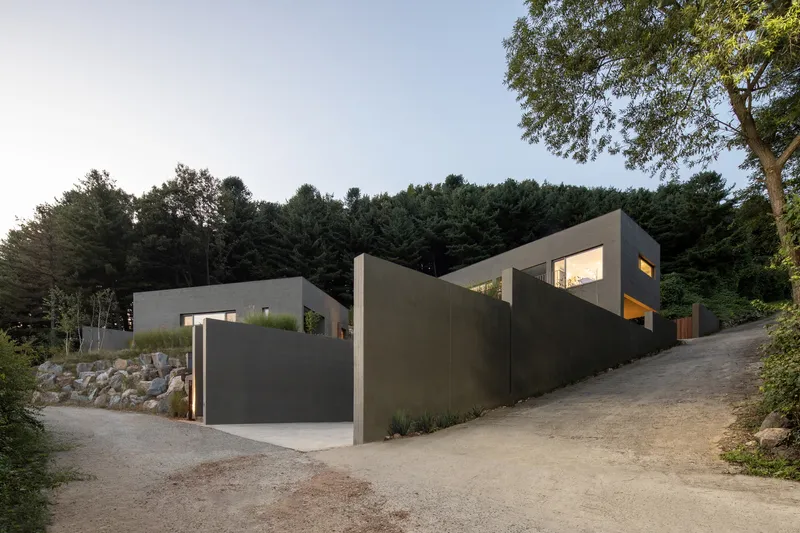Street view of clustered concrete volumes with illuminated windows nestled into a wooded hillside at twilight