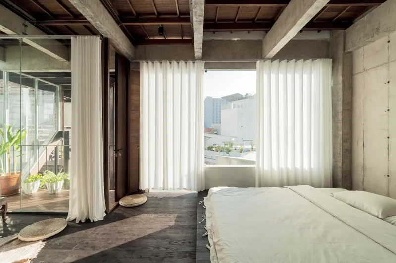 Bedroom with exposed timber ceiling, concrete columns, and sheer curtains framing the view to adjacent buildings