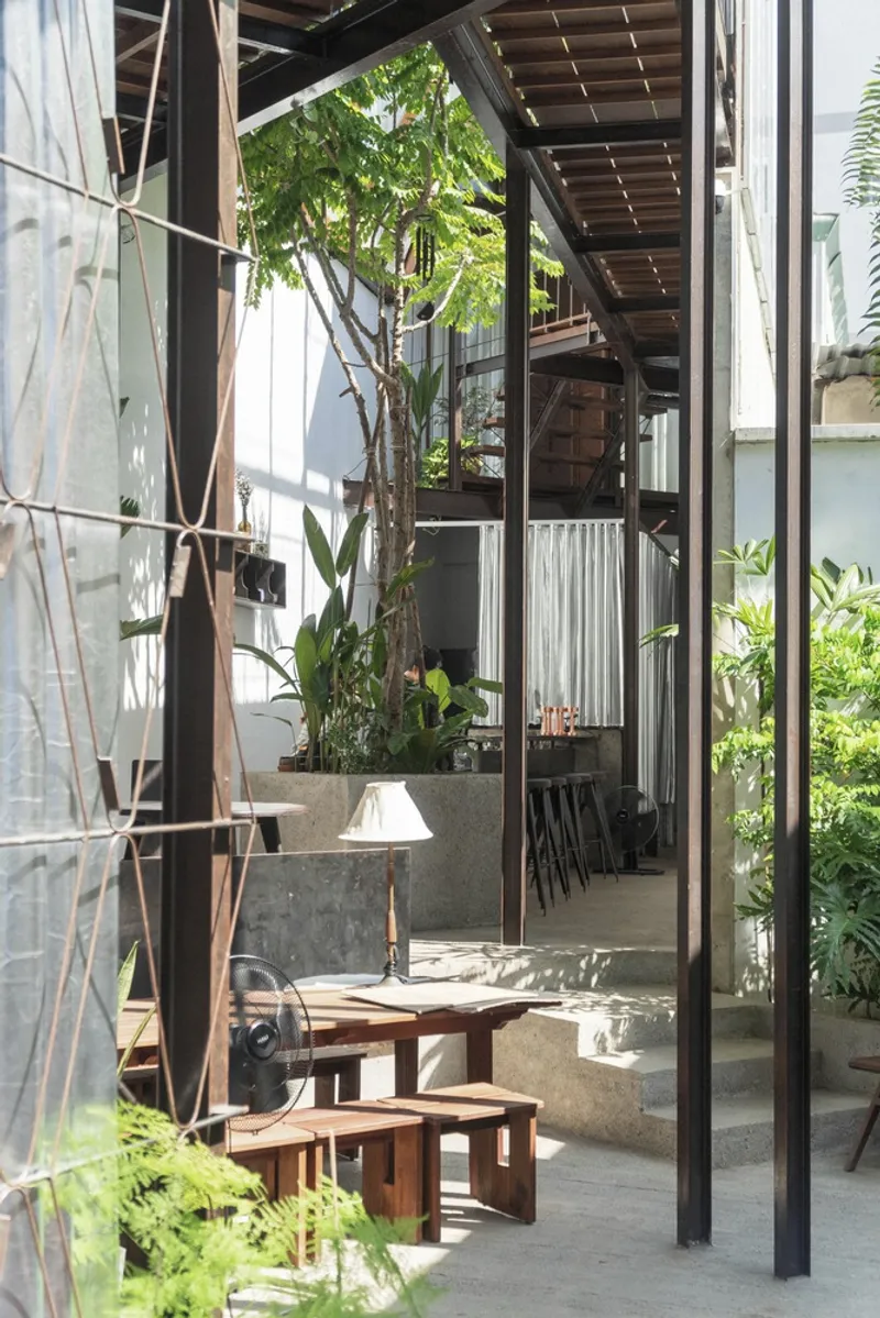Steel frame structure with timber roof grid above a concrete planter and timber furniture in dappled sunlight