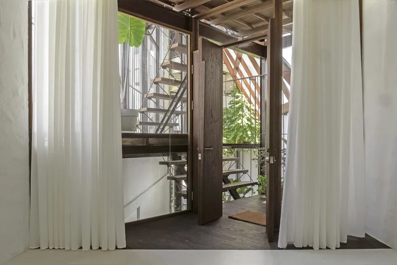 Open pivot door framed by white curtains revealing an outdoor steel staircase and timber beams overhead