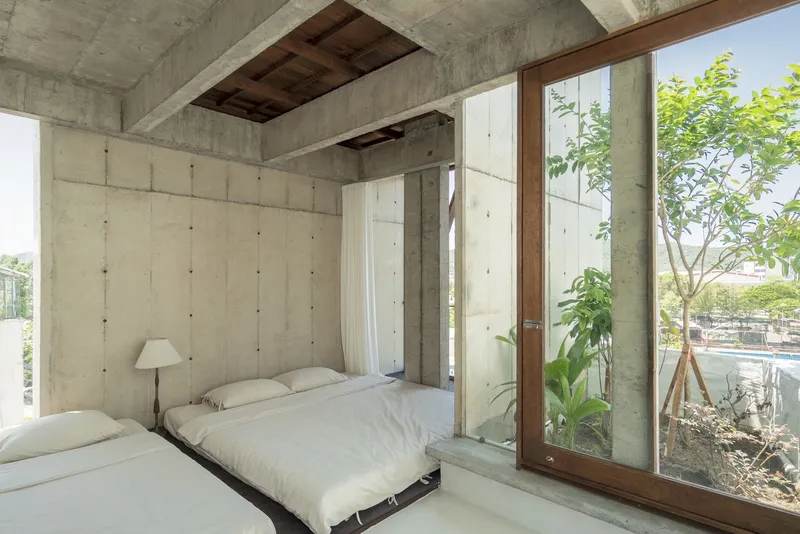 Bedroom with exposed concrete walls and ceiling opening to a planted courtyard through timber framed glazing