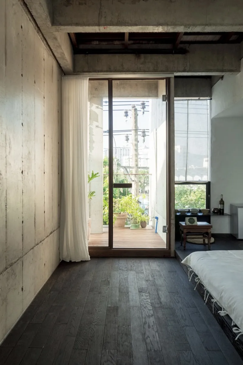 Bedroom with board-formed concrete walls, dark timber flooring, and glazed doors opening to a planted balcony