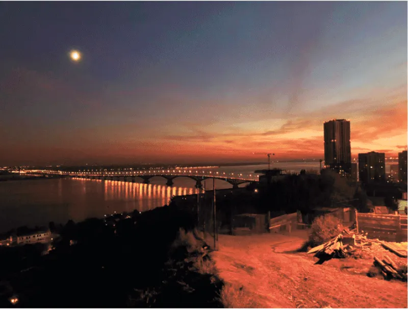 View across a river toward an illuminated bridge and city towers at dusk with moon above