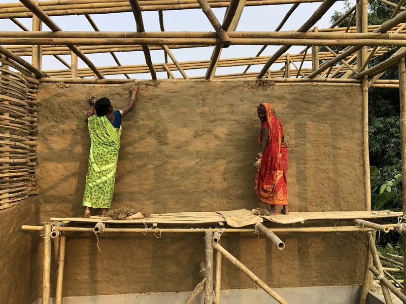 Two workers applying earth plaster to an interior wall beneath the bamboo roof framework
