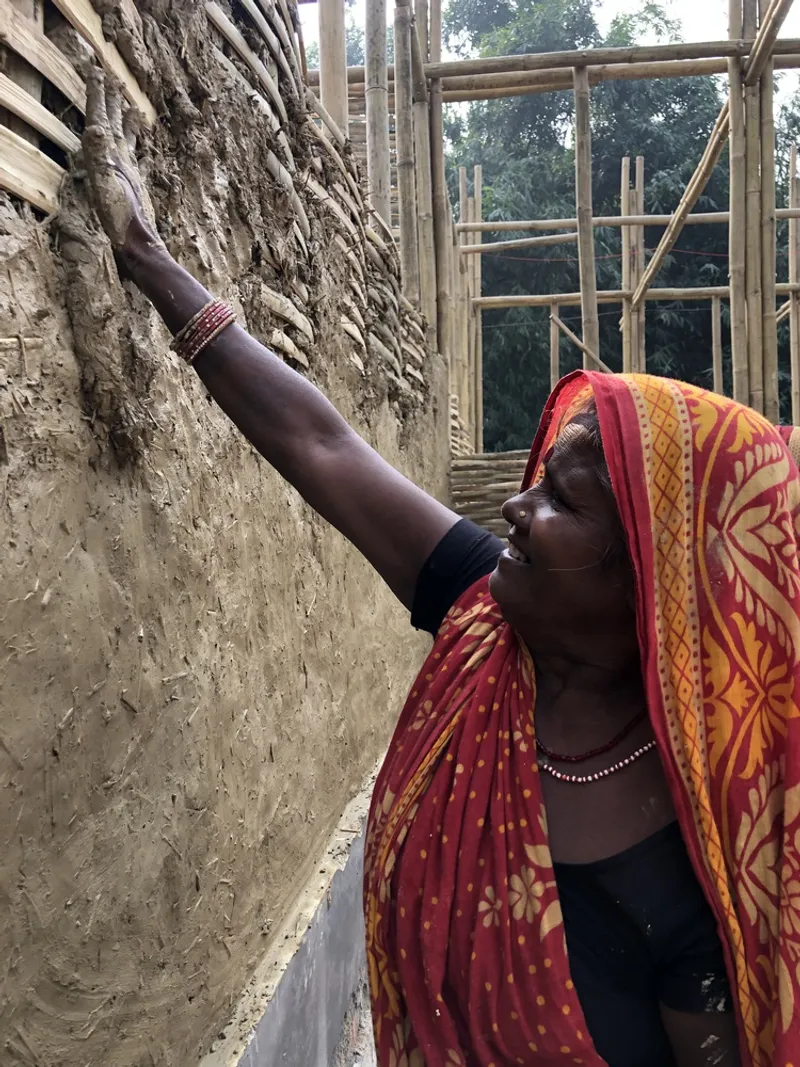 Woman in patterned garment applying clay and straw mixture to woven bamboo wall during construction