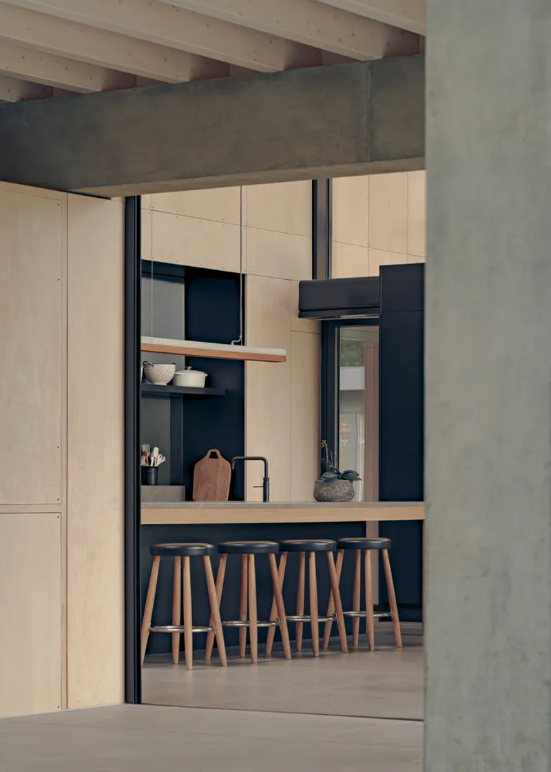 Kitchen with timber bar stools and pale plywood cabinetry framed by concrete column