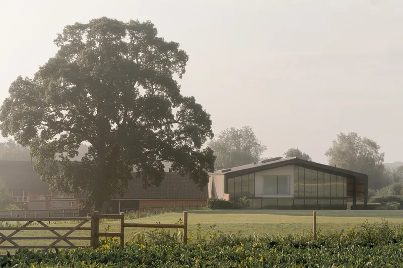 Long view of the low-slung pavilion with barrel roof and glazed facade across a mown lawn in morning mist