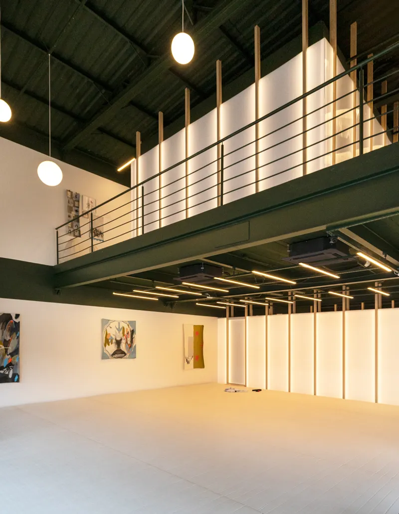 Double-height gallery space with exposed green ceiling structure and suspended mezzanine with metal railing