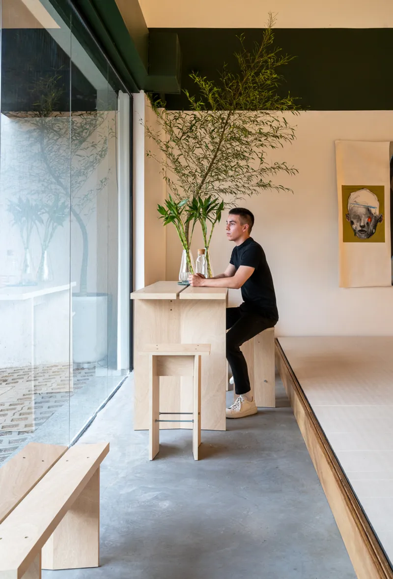 Person at plywood bar-height table with vase of bamboo beside floor-to-ceiling glazing overlooking courtyard