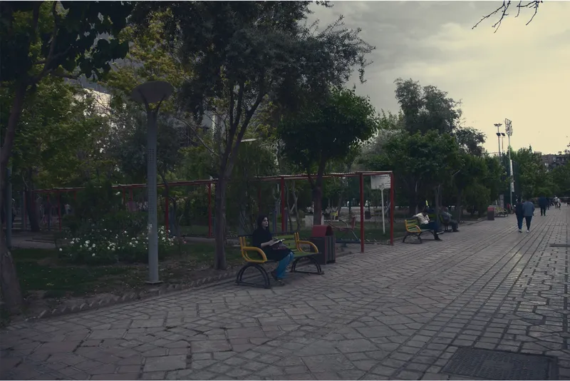 Paved plaza with red metal playground equipment and yellow benches beneath mature trees