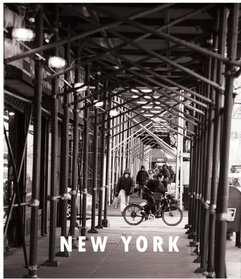 Covered pedestrian passage with exposed steel truss ceiling and diagonal columns as a cyclist passes through
