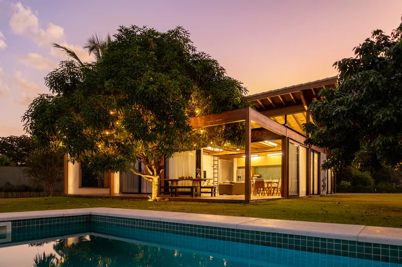 Garden pavilion with timber pergola and exposed roof structure illuminated at dusk beside the pool
