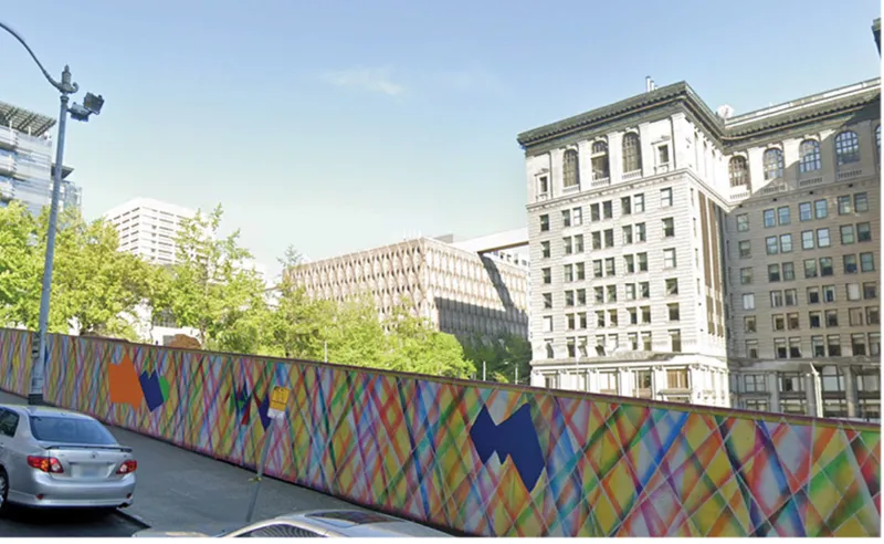 Construction fence with diagonal multicolored stripe pattern along a street with trees and passing traffic