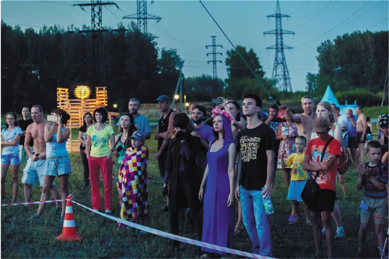 Crowd gathered at dusk near transmission towers and a glowing timber installation in an open field