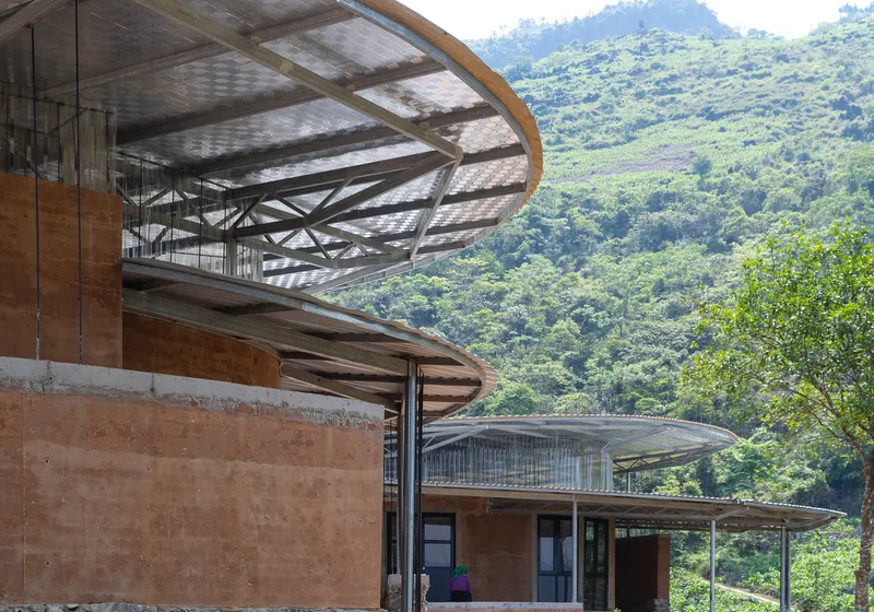 Cantilevered circular roof canopy with exposed steel structure above rammed earth walls against a green hillside