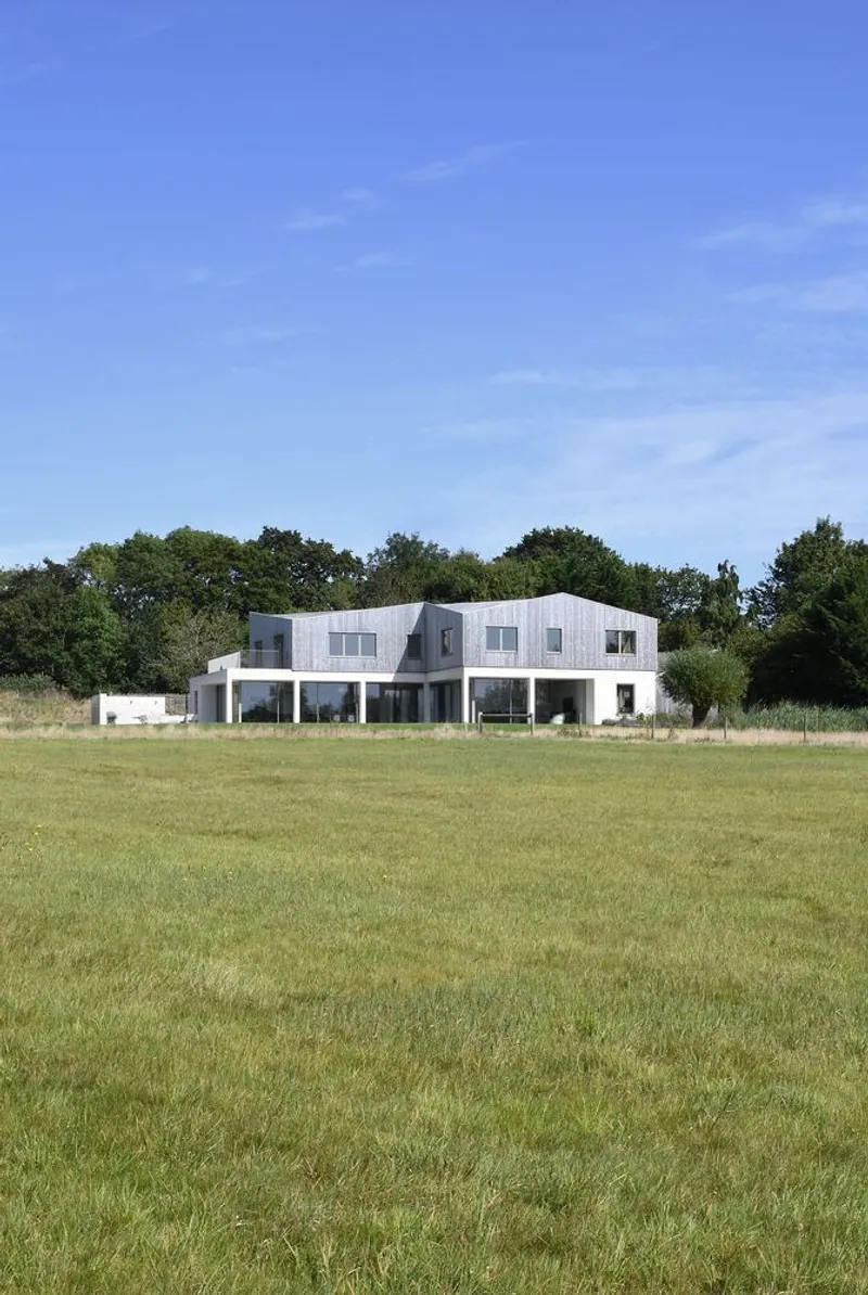Rear elevation showing white rendered facade with upper shingle cladding across a mown meadow under blue sky
