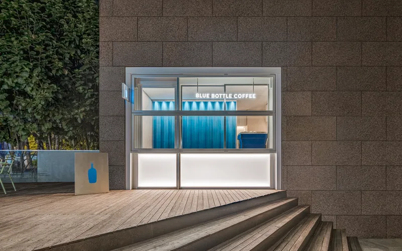 Glass-fronted kiosk with blue corrugated metal wall and timber deck steps at dusk